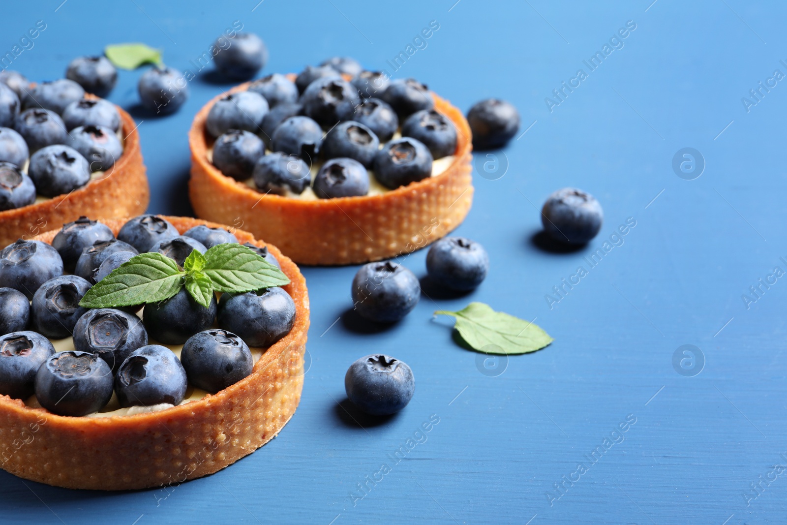 Photo of Tartlets with fresh blueberries and mint on blue table, space for text. Delicious dessert