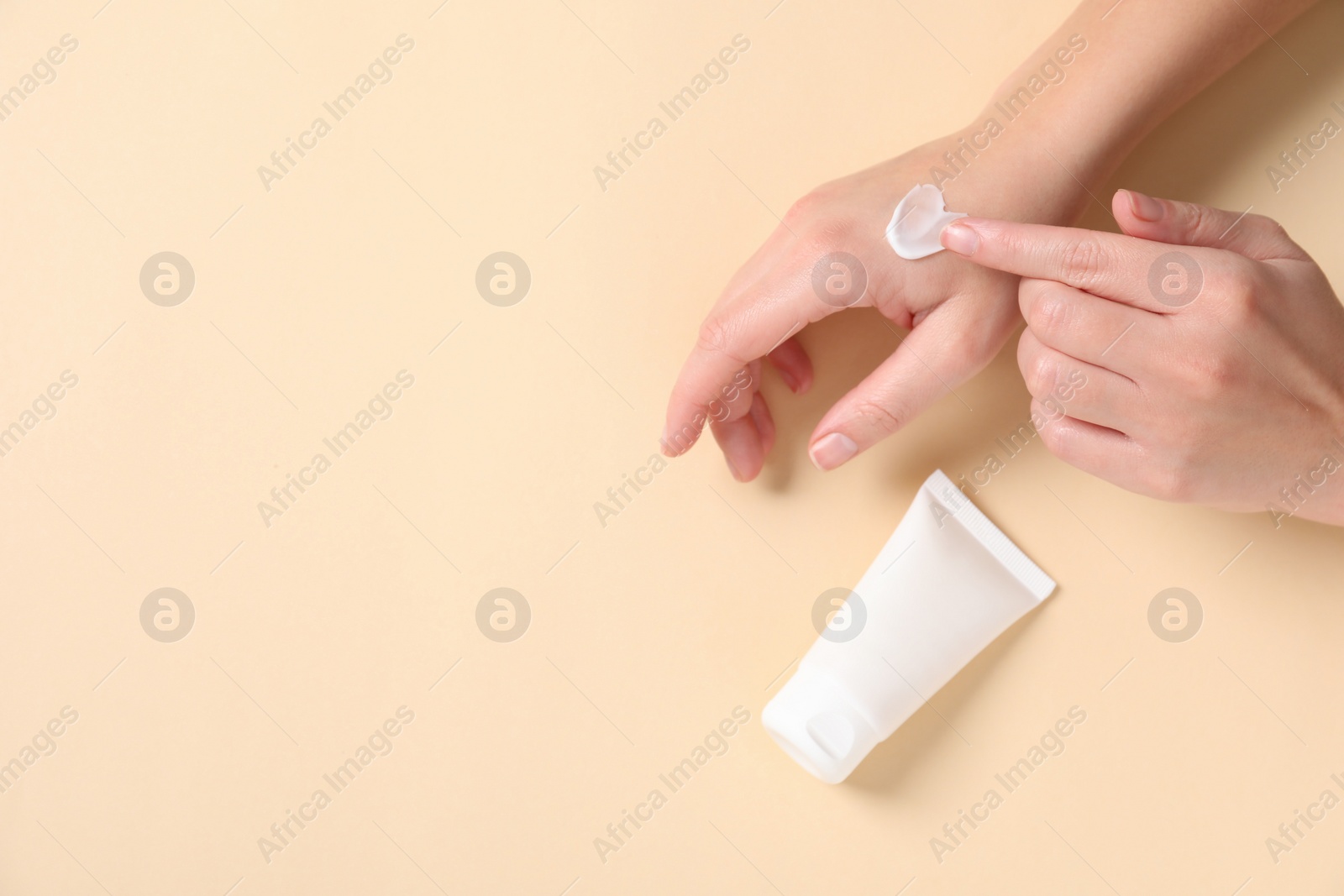 Photo of Woman with tube applying cosmetic cream onto her hand on beige background, top view. Space for text