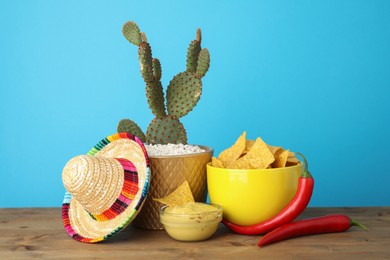 Photo of Mexican sombrero hat, cactus, chili peppers, nachos chips and guacamole in bowls on wooden table against light blue background