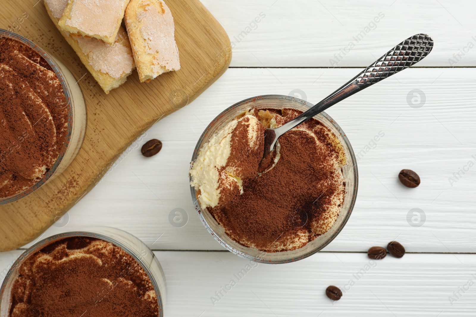 Photo of Delicious tiramisu in glasses, spoon, coffee beans and cookies on white wooden table, top view