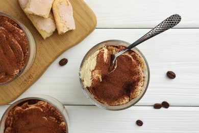 Photo of Delicious tiramisu in glasses, spoon, coffee beans and cookies on white wooden table, top view