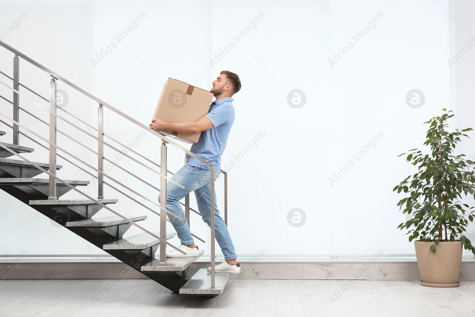 Photo of Young man carrying carton box upstairs indoors. Posture concept