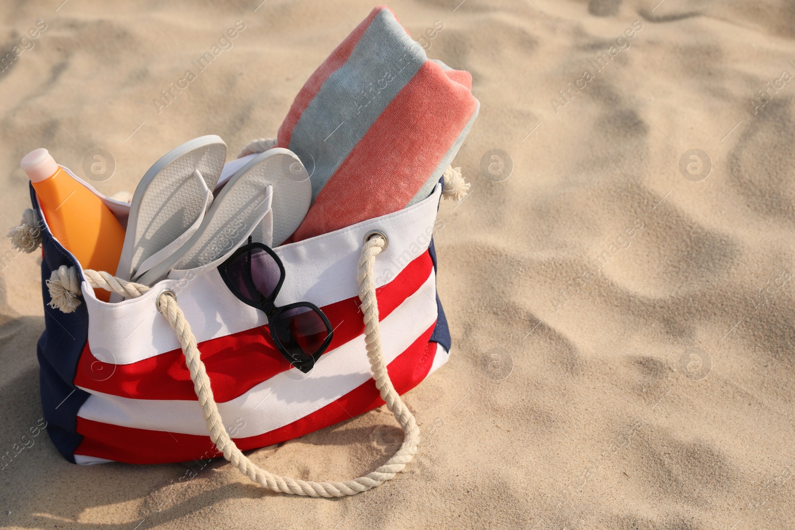 Photo of Beach bag with flip flops, towel, sunglasses and sunscreen on sand, space for text