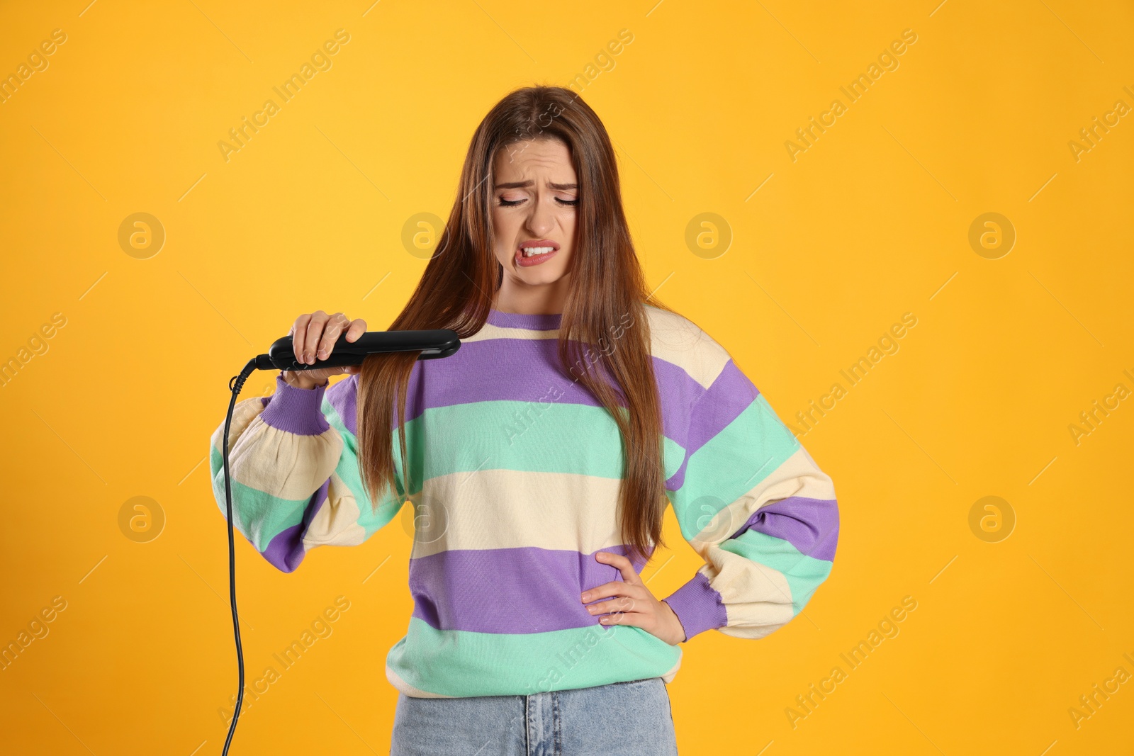 Photo of Upset young woman with flattening iron on yellow background. Hair damage