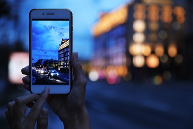 Photo of Woman taking photo of modern city in evening with smartphone, focus on hand. Bokeh effect