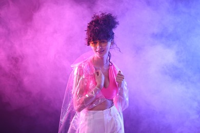 Photo of Beautiful young woman in transparent coat and sunglasses posing on color background in neon lights and smoke