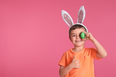 Photo of Little boy in bunny ears headband holding Easter egg near eye on color background, space for text