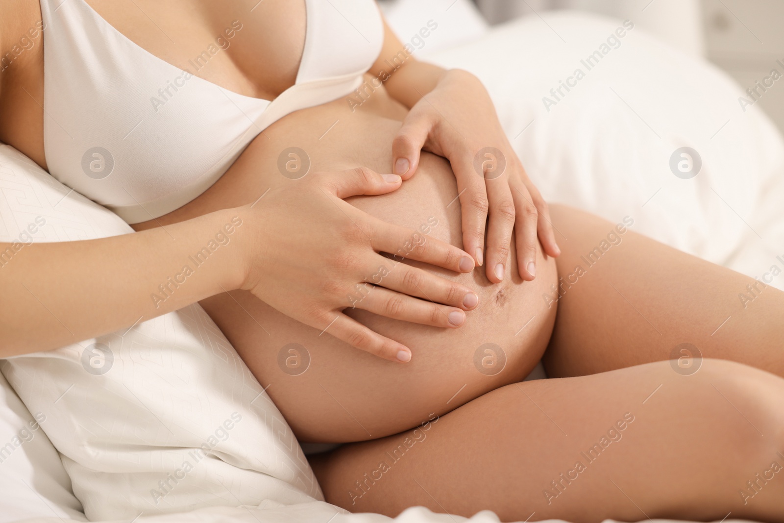 Photo of Pregnant woman in stylish comfortable underwear making heart with hands on her belly on bed, closeup