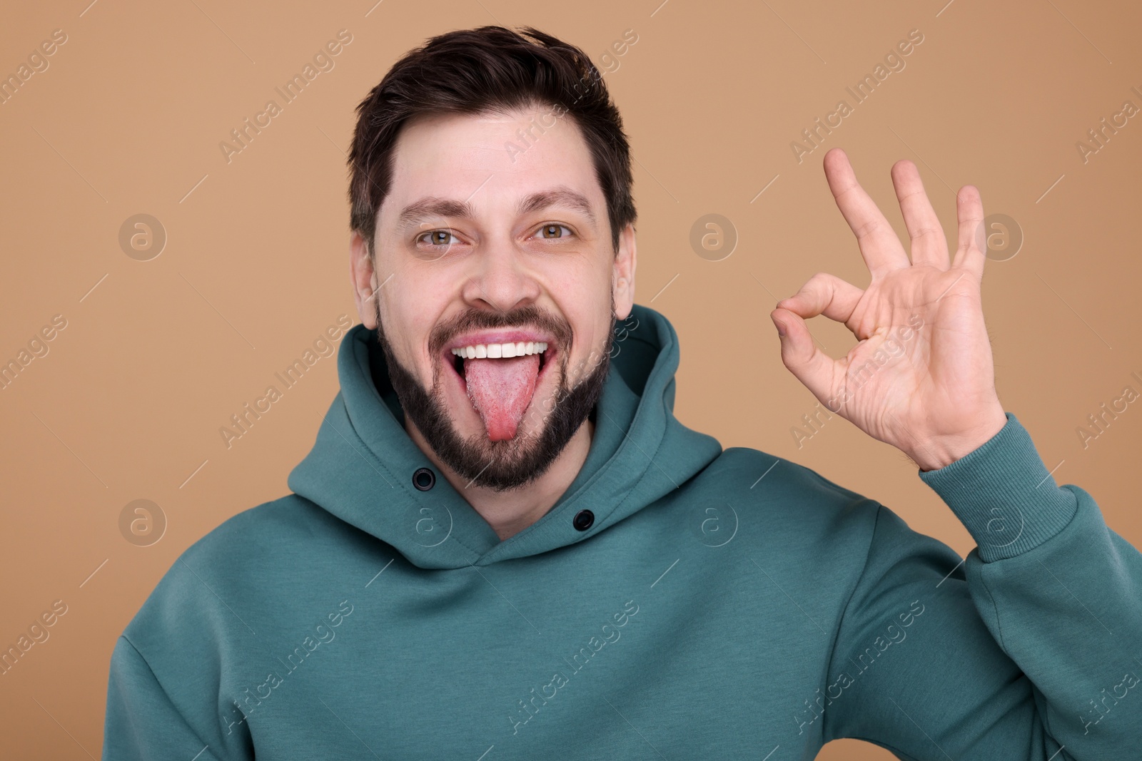 Photo of Happy man showing his tongue and making ok gesture on beige background