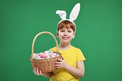 Photo of Easter celebration. Cute little boy with bunny ears and wicker basket full of painted eggs on green background