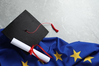 Photo of Black graduation cap, diploma and flag of European Union on light grey marble table, flat lay. Space for text