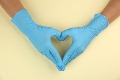 Person in medical gloves making heart with hands on beige background, top view