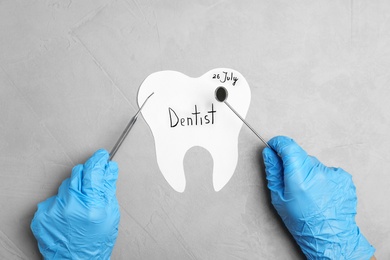 Dentist holding professional tools over paper tooth on grey background, top view