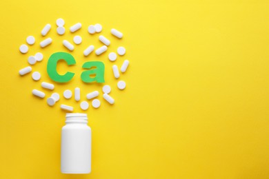 Photo of Pills, open bottle and calcium symbol made of green letters on yellow background, flat lay. Space for text