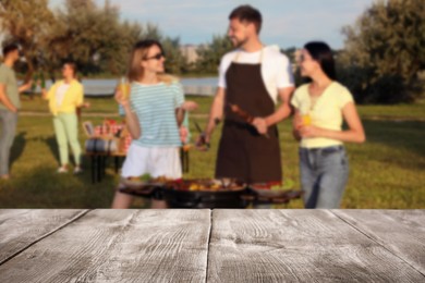Image of Empty wooden table and blurred view of friends cooking food on barbecue grill in park
