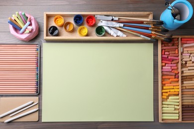 Photo of Blank sheet of paper, colorful chalk pastels and other drawing tools on wooden table, flat lay. Modern artist's workplace