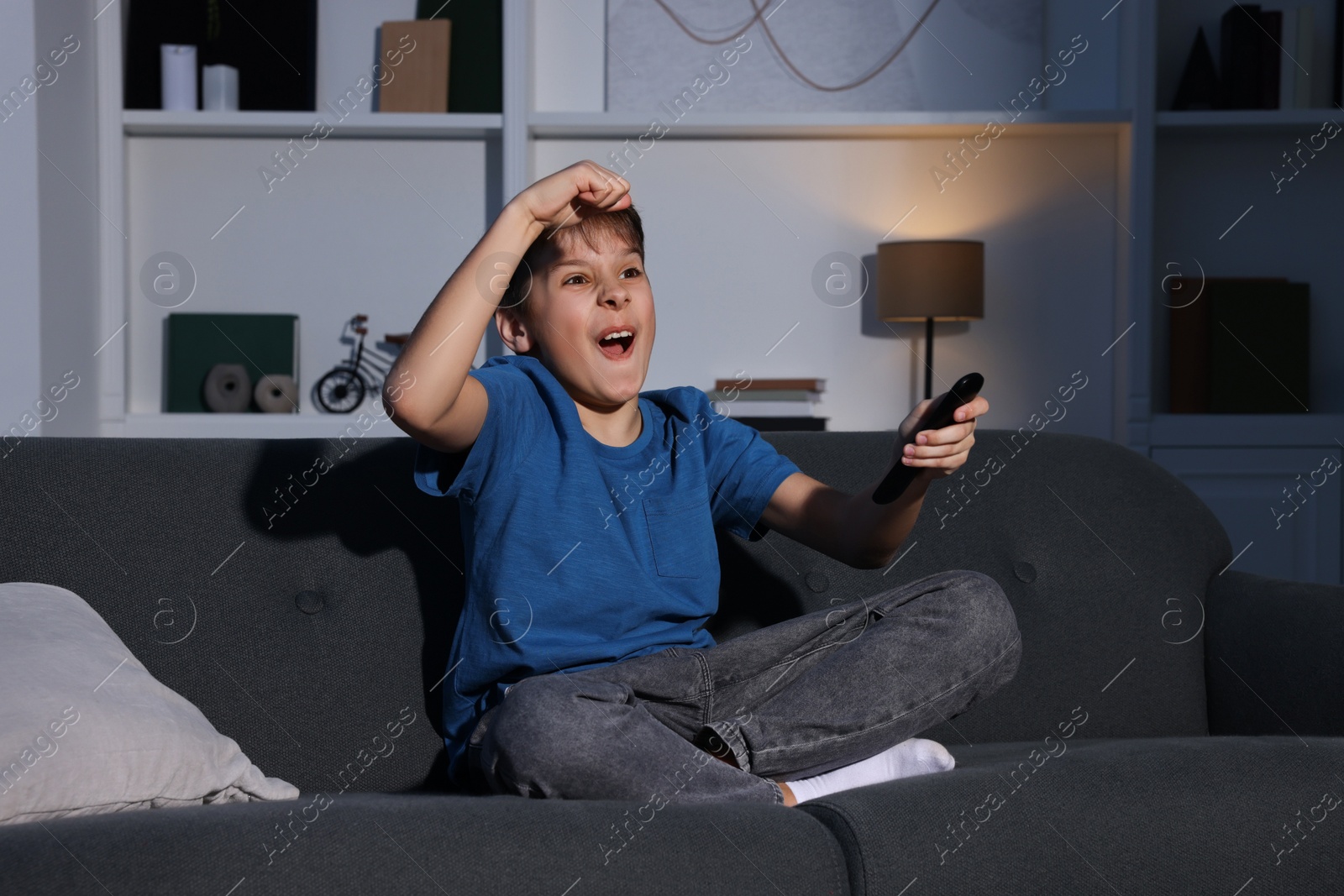Photo of Emotional boy watching TV and holding remote control on sofa at home