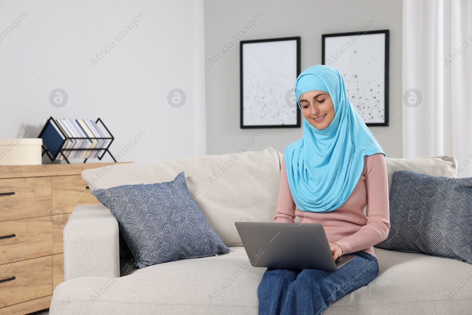 Photo of Muslim woman using laptop at couch in room. Space for text