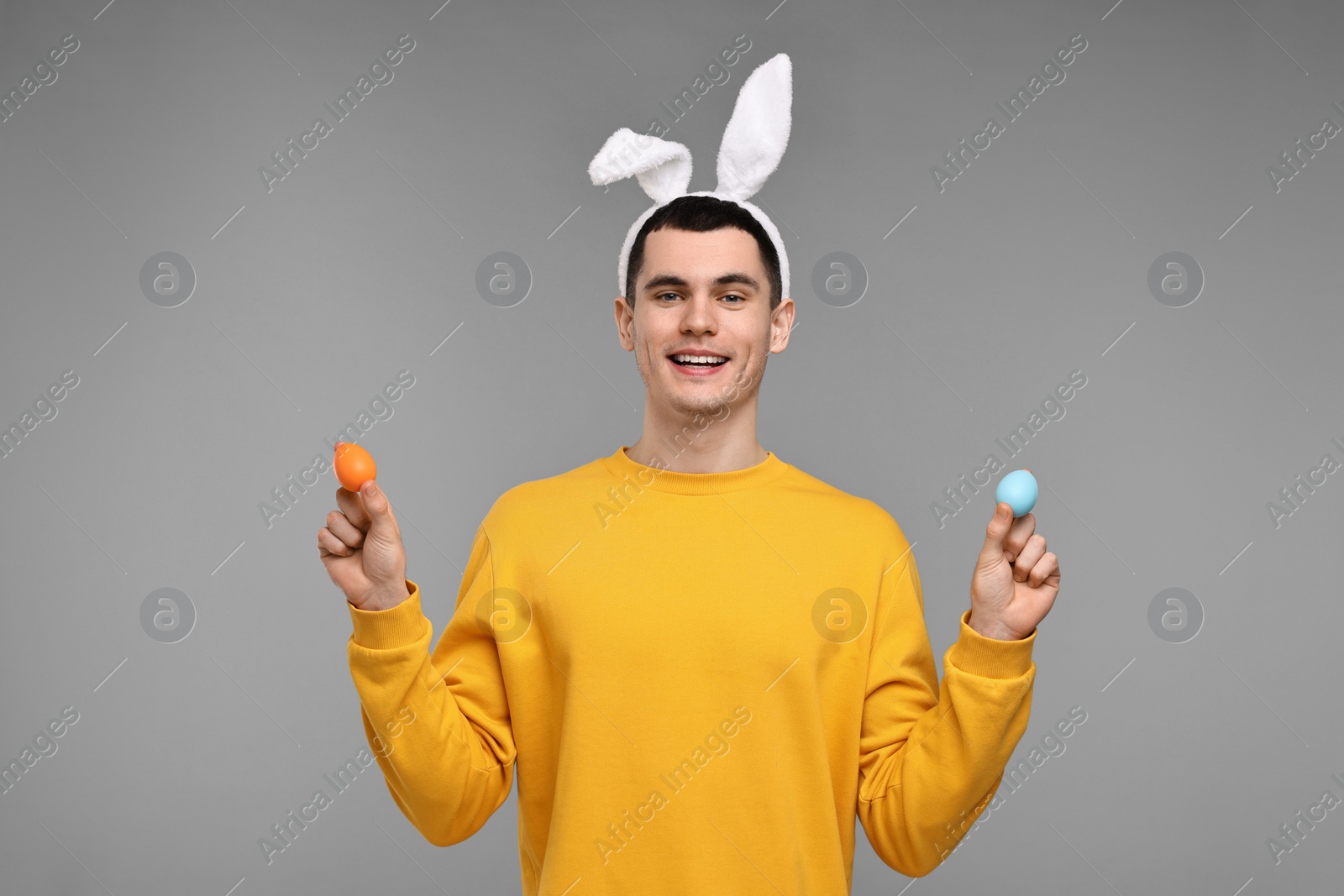 Photo of Easter celebration. Handsome young man with bunny ears holding painted eggs on grey background