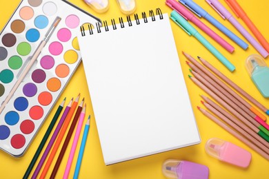 Watercolor palette, empty notebook, colorful pencils and markers on yellow background, flat lay. Space for text