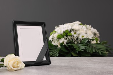 Photo of Photo frame with black ribbon, rose on light table and wreath of flowers near grey wall indoors. Funeral attributes