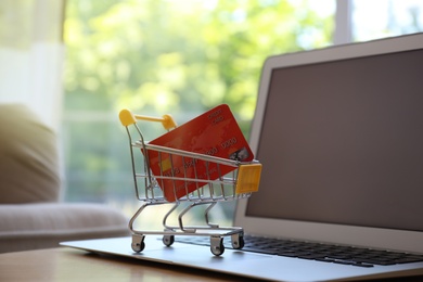 Photo of Internet shopping. Laptop and small cart with credit card on table indoors
