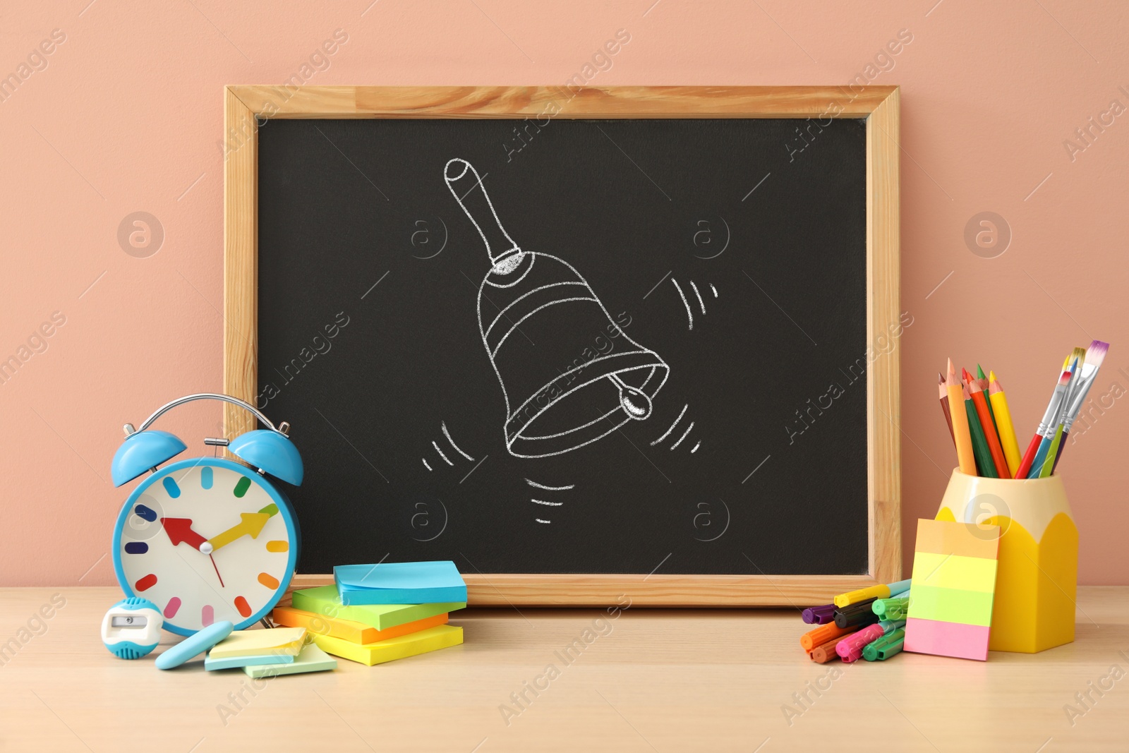 Image of Small blackboard with drawn school bell and different stationery on wooden table near pink wall 