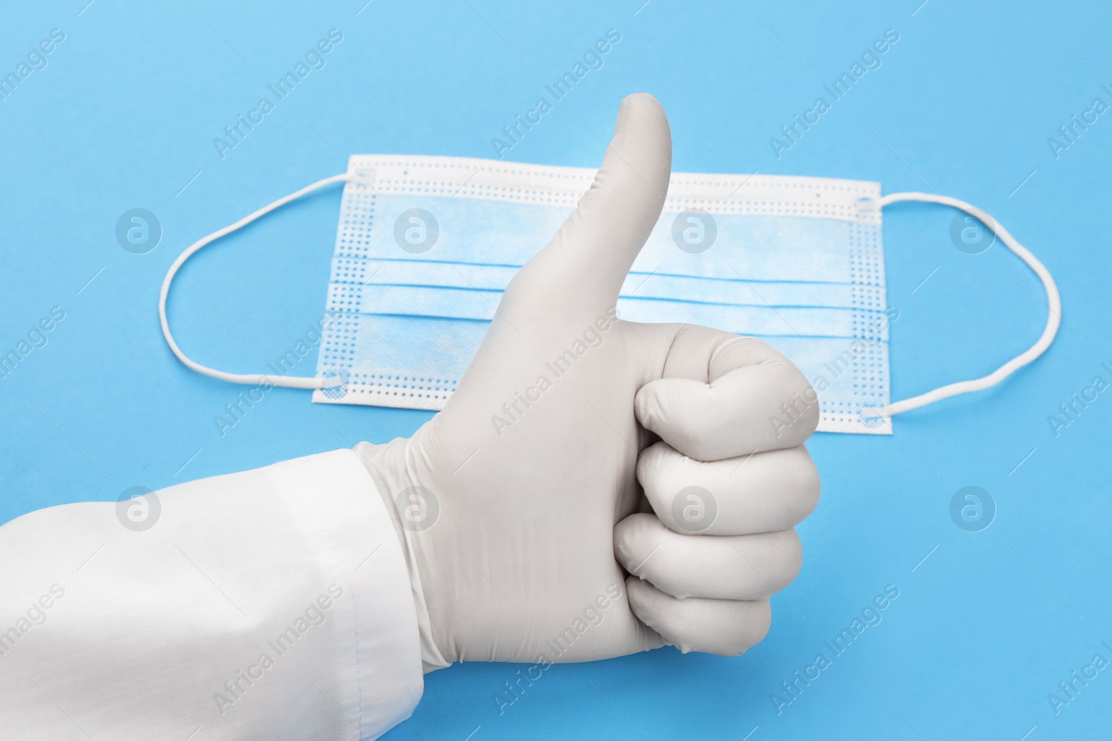 Photo of Doctor in medical gloves showing thumb up gesture near protective mask on light blue background, closeup