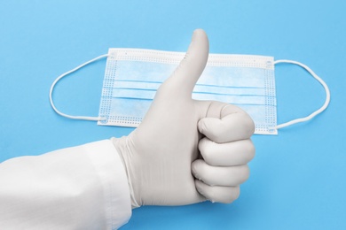 Photo of Doctor in medical gloves showing thumb up gesture near protective mask on light blue background, closeup