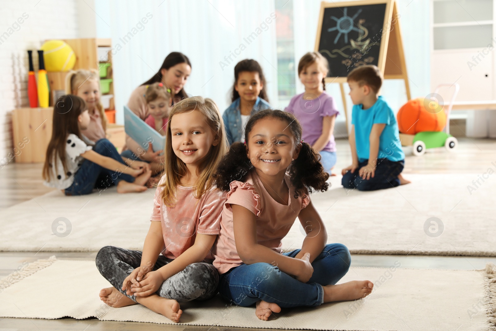 Photo of Cute girls sitting on floor while kindergarten teacher reading book to other children indoors