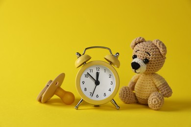 Alarm clock, toy bear and baby dummy on yellow background. Time to give birth