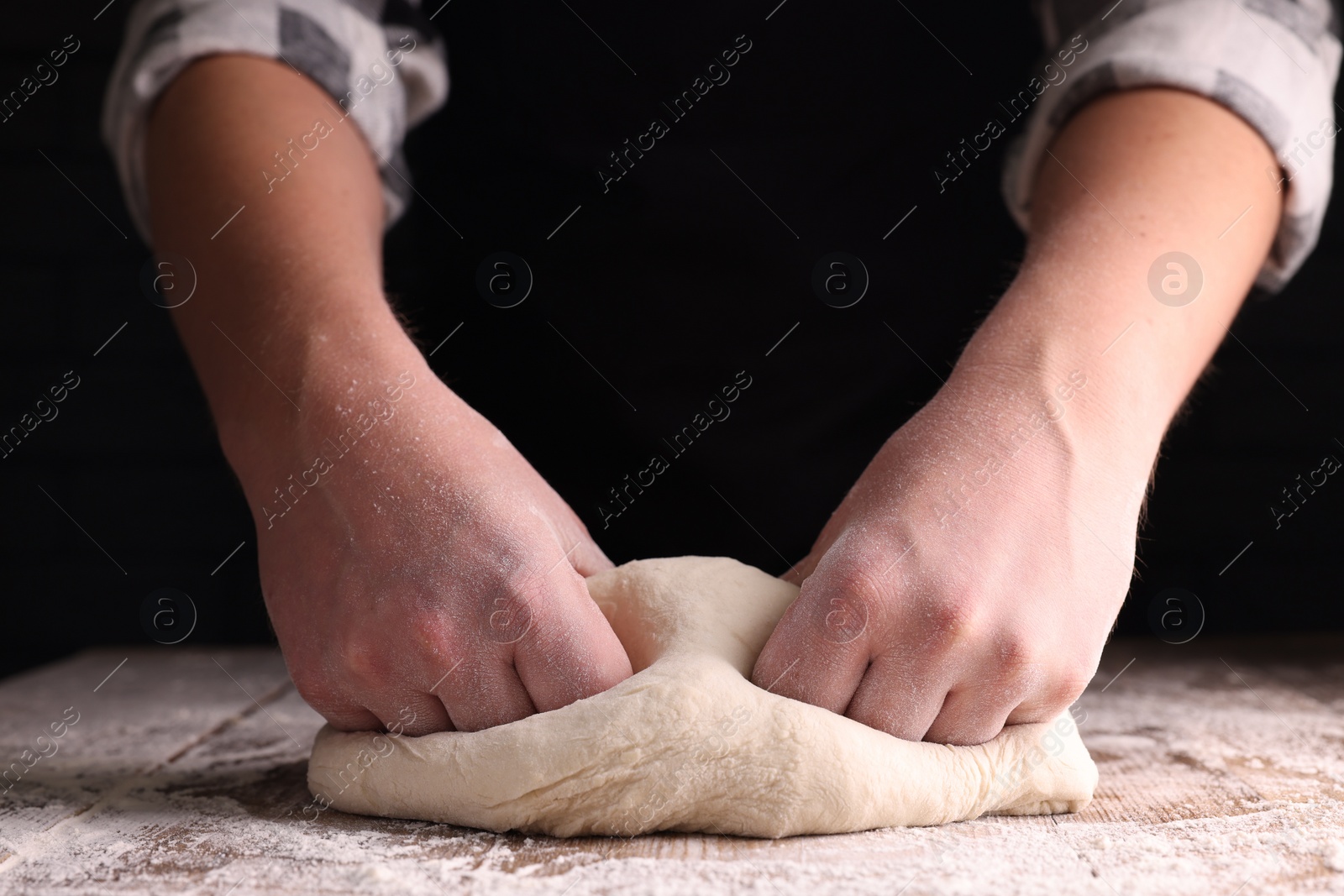 Photo of Man kneading dough at wooden table on dark background, closeup