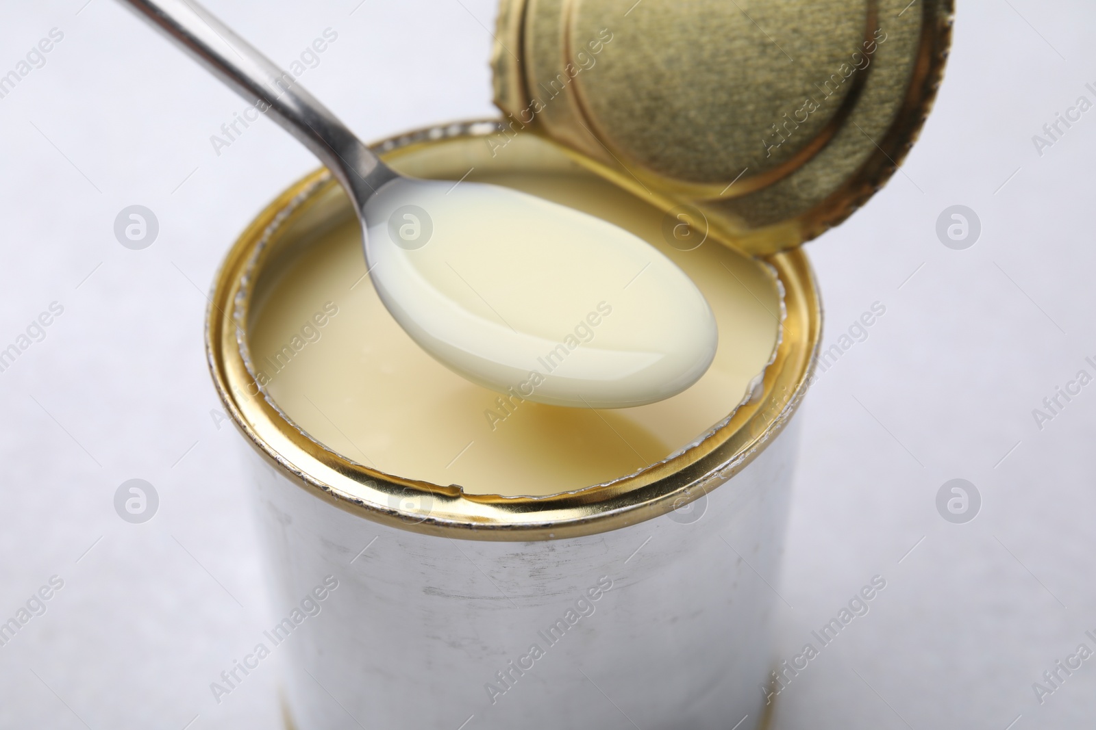 Photo of Spoon with condensed milk over open tin can on white table, closeup