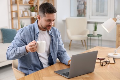 Photo of Man with cup of drink working on laptop at wooden desk in room