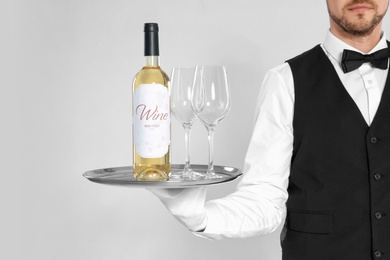 Young waiter holding tray with glasses and bottle of wine on light background