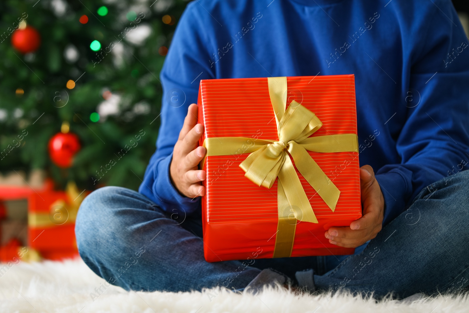 Photo of Man in blue sweater with Christmas gift sitting on fluffy carpet at home, closeup