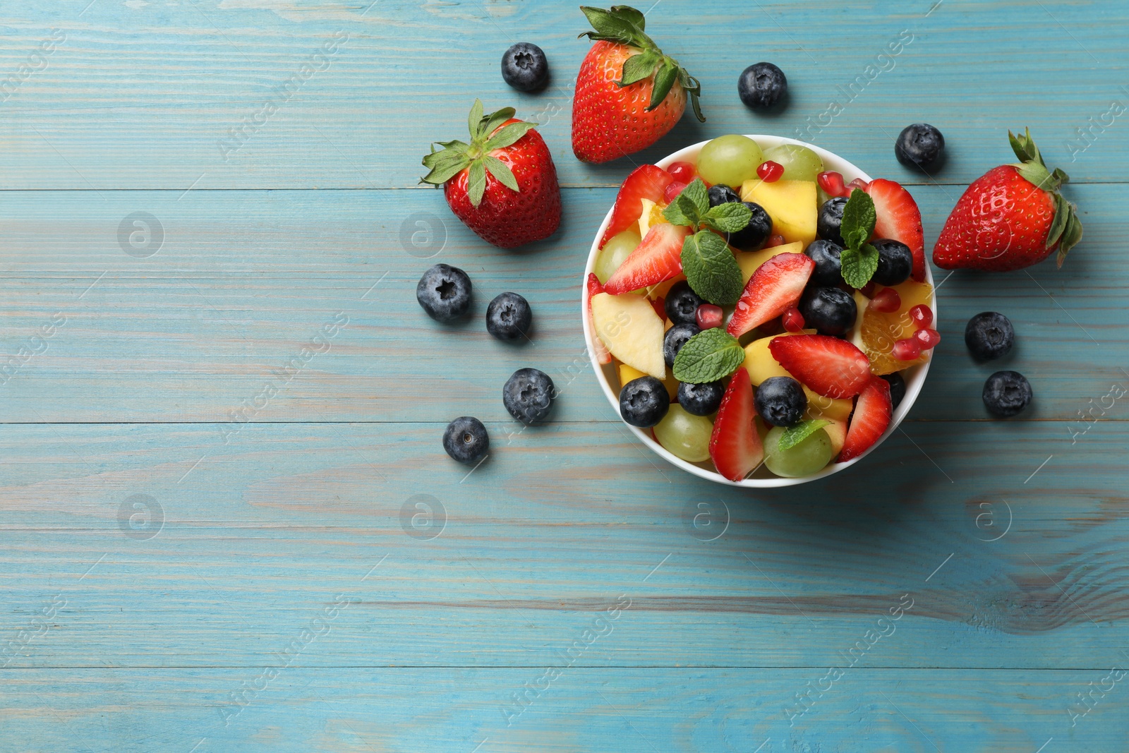 Photo of Tasty fruit salad in bowl and ingredients on light blue wooden table, flat lay. Space for text