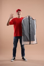 Photo of Dry-cleaning delivery. Happy courier holding garment cover with clothes and showing OK gesture on beige background