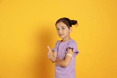Photo of Vaccinated little girl with medical plaster on her arm showing thumb up against yellow background