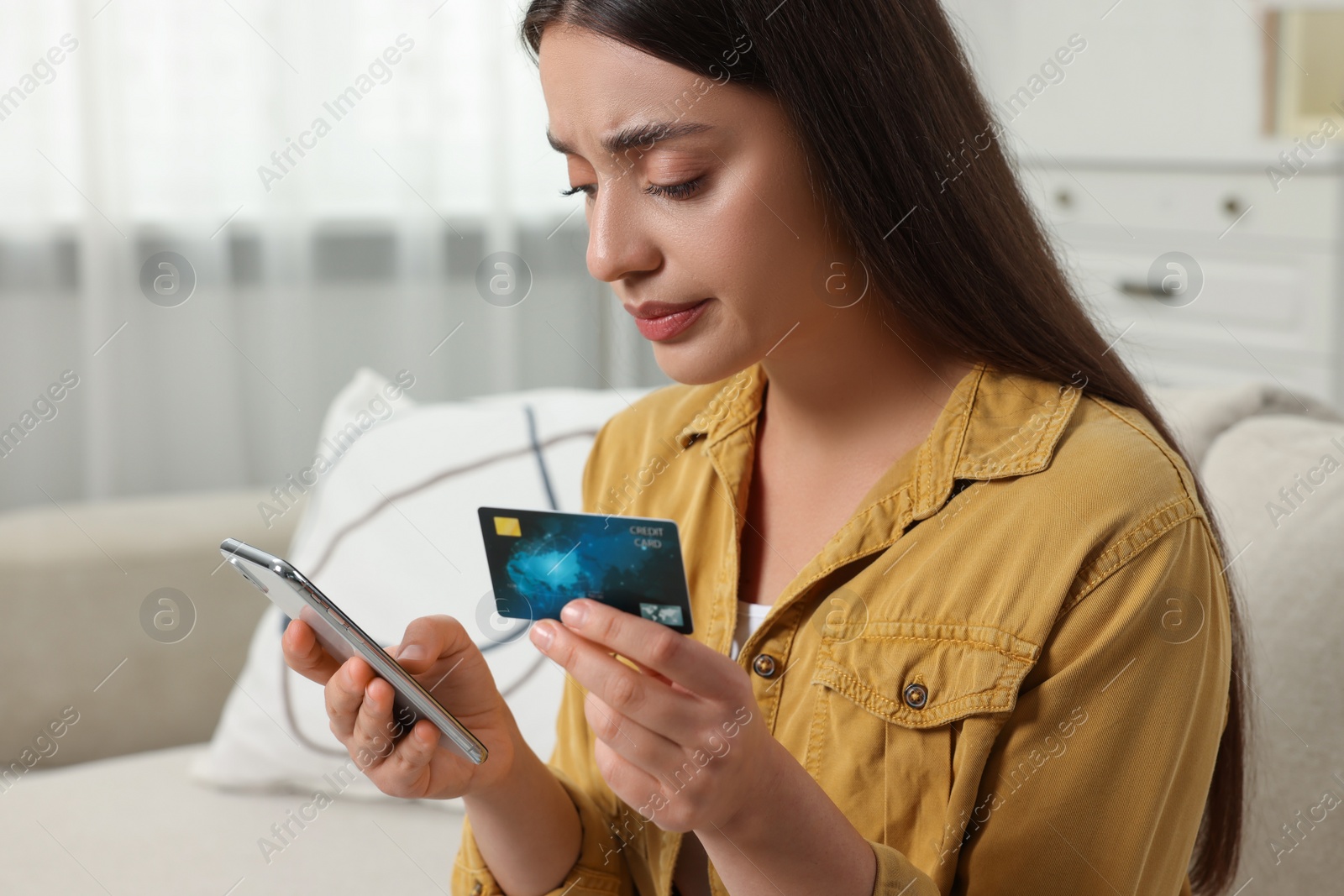 Photo of Confused woman with credit card using smartphone at home. Be careful - fraud