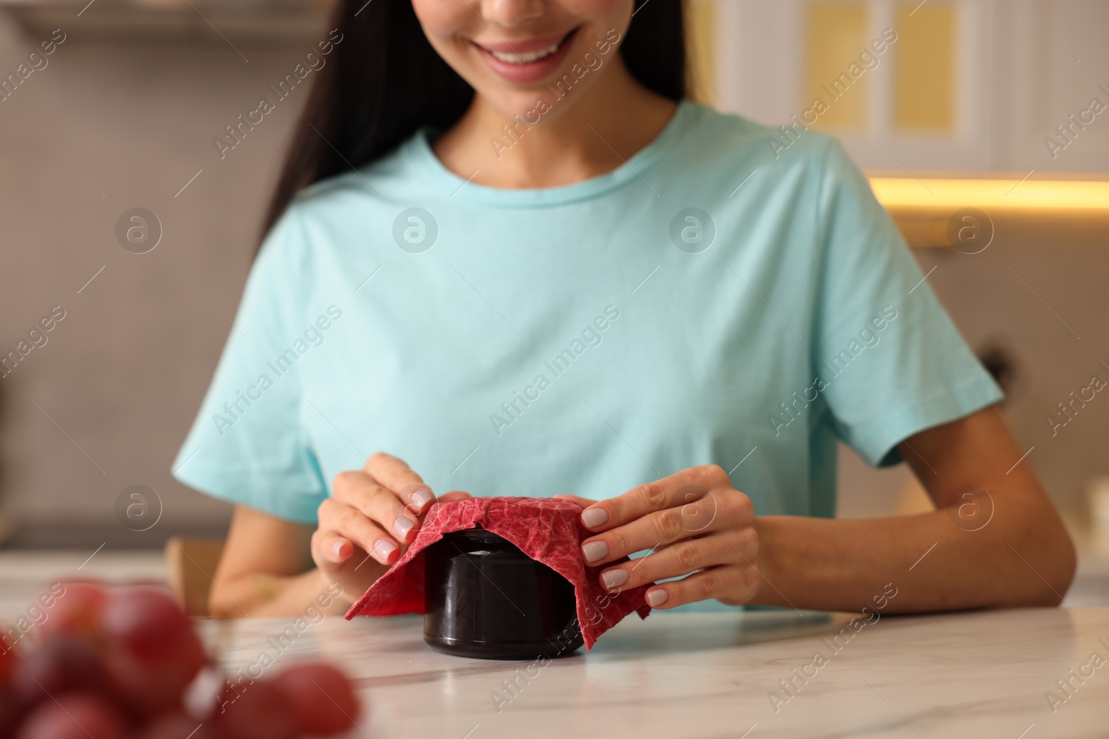 Photo of Woman packing jar of jam into beeswax food wrap at light table in kitchen, closeup