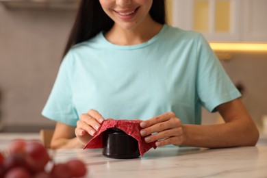 Woman packing jar of jam into beeswax food wrap at light table in kitchen, closeup