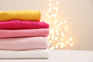 Photo of Stack of folded clothes on wooden table against beige background with blurred lights, space for text