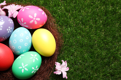 Photo of Colorful Easter eggs in decorative nest on green grass, top view. Space for text