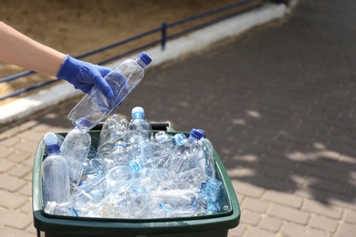 Photo of Woman in gloves putting used plastic bottle into trash bin outdoors, closeup. Recycling problem