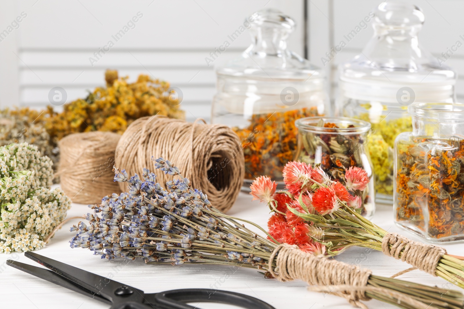 Photo of Bunches of dry flowers, different medicinal herbs, scissors and spools on white wooden table