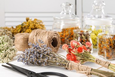 Photo of Bunches of dry flowers, different medicinal herbs, scissors and spools on white wooden table