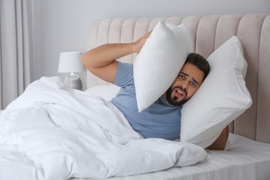 Photo of Unhappy young man covering ears with pillows in bed at home. Noisy neighbours