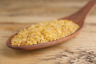 Photo of Spoon with uncooked bulgur on wooden table, closeup