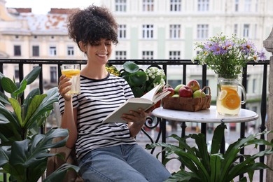 Photo of Young woman with refreshing drink reading book near beautiful houseplants on balcony